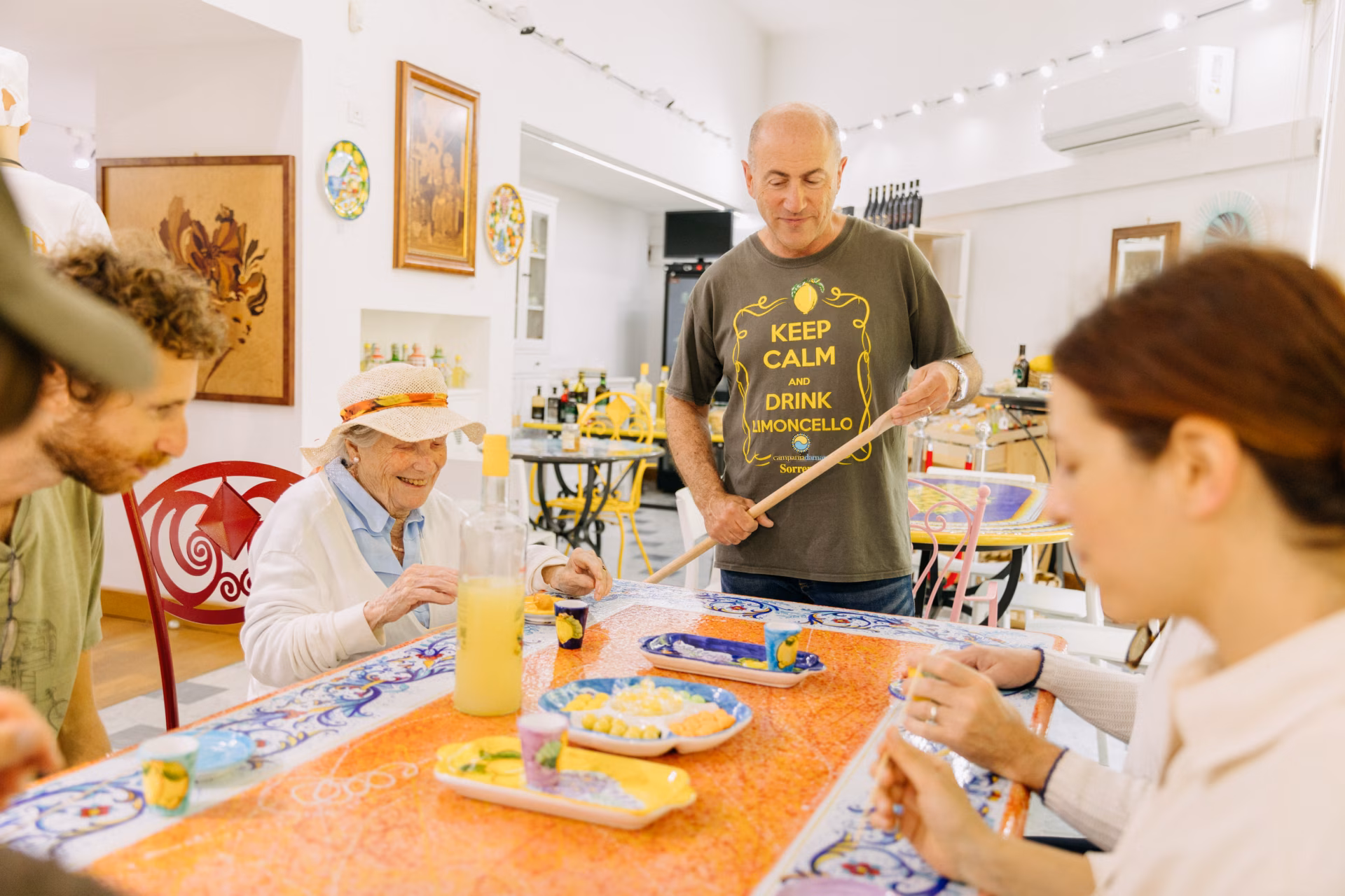 A local host wearing a "Keep Calm and Drink Limoncello" shirt pouring fresh lemon liqueur into colorful ceramic glasses for guests seated around a decorative majolica tile table during an Eating Europe food tour.